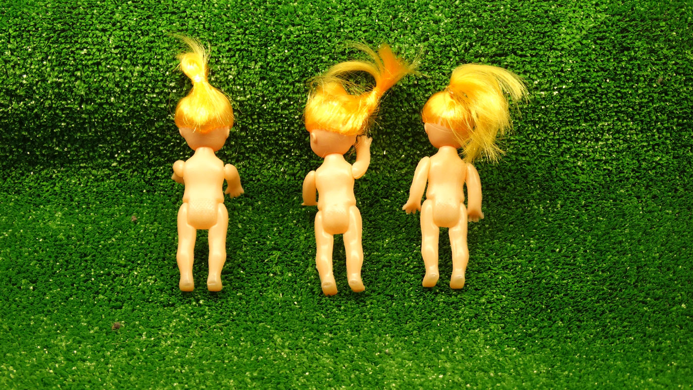 Plakat ze spektaklu Czekając trawnika. Na zielonym tle plakatu imitujące trawnik leżą na brzuchu trzy gołe lalki. Wszystkie mają żółte włosy spięte w kucyk.