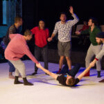 Zdjęcie ze spektaklu Czytanki performatywne. Na scenie znajduje się sześciu tancerzy w kolorowych dresach. Jeden z nich leży na ziemi a reszta stoi w kole wokół niego