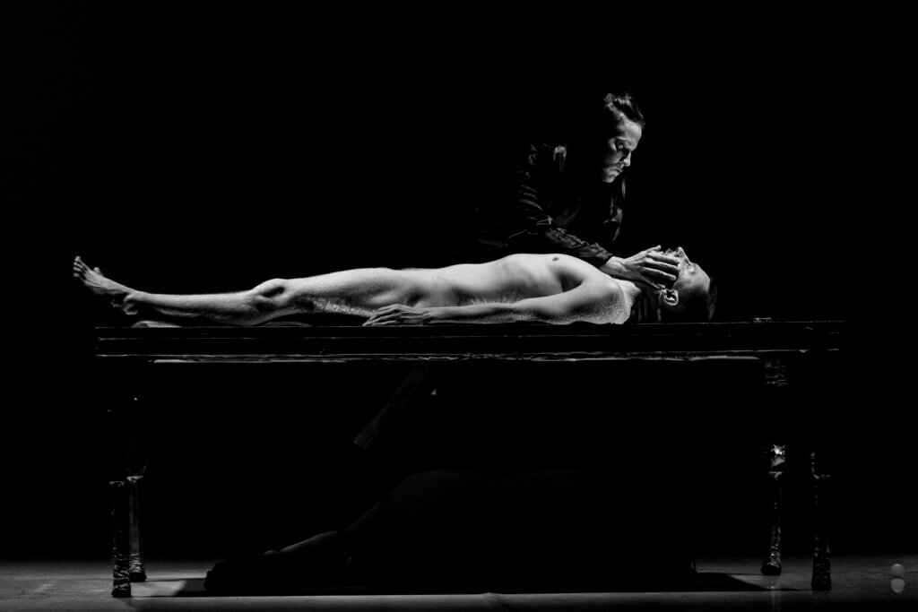 Zdjęcie czarno-białe ze spektaklu Panny z przyszłośći. Na zdjęciu znajduje się nagi męzczyzna leżący na plecach na stole a nad nim pochyla się kobieta, jej prawa ręka dotyka twarzy mężczyzny.