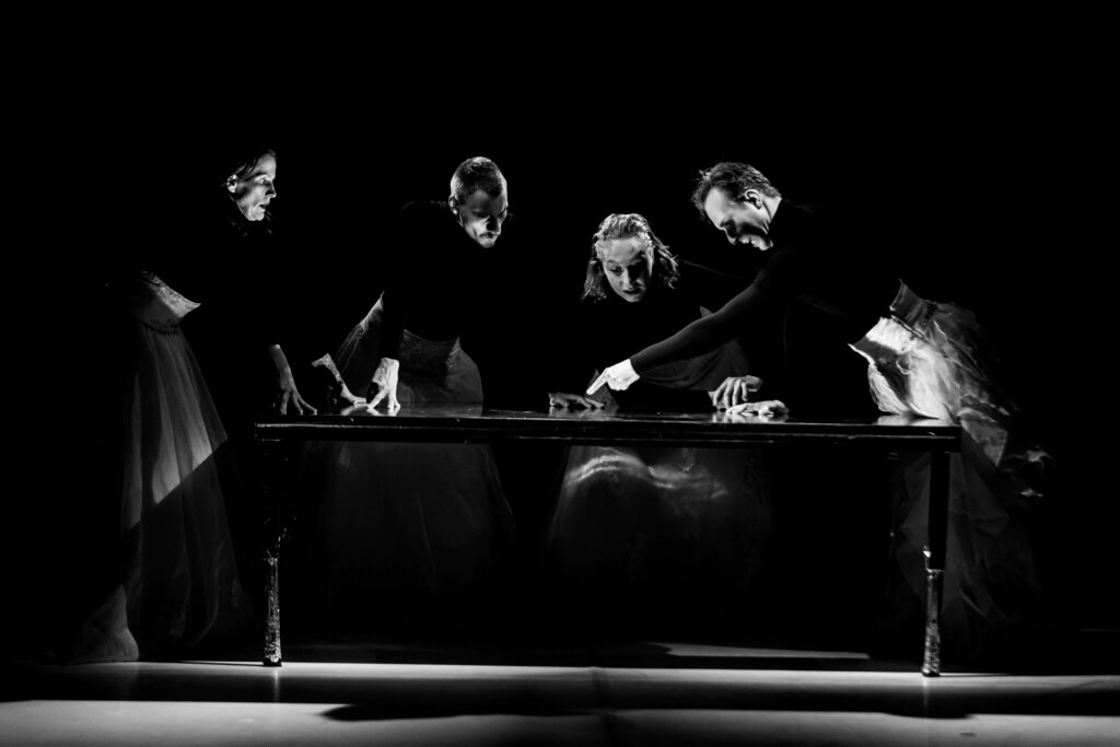 Zdjęcie ze spektaklu Panny z przyszłości. na zdjeciu znajdują się cztery osoby wokół prostokątnego stołu. Trzy osoby wspierają się rękoma wzrok mają skierowany na palec meżczyzny, który coś wskazuje na stole. Wszyscy ubrani są w czarne koszulki z długim rękawem oraz białe długie spódnice. Zdjęcie jest czarno-białe