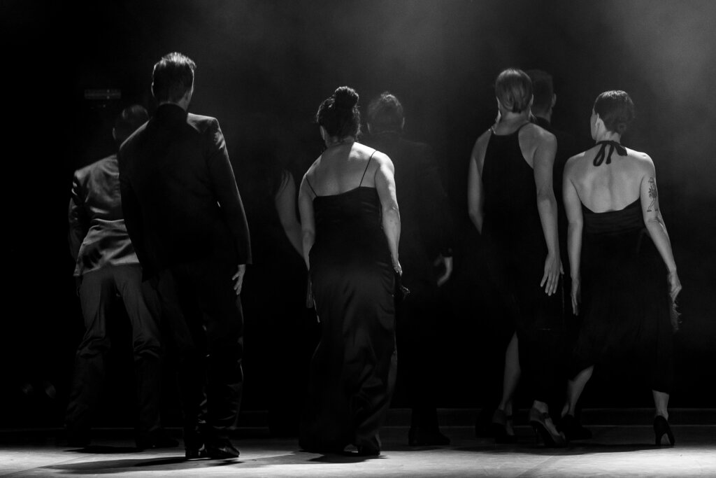 Tribule To zdjęcie czarno białe. Na zdjęciu znajdują się tancerze obróceni tyłem do publiczności, z lewej strony dwóch mężczyzn ubranych w garnitury a po prawej stronie znajduje się kobiety ubrane w wieczorowe suknie.