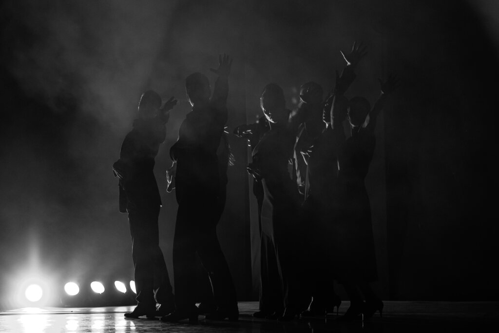 Zdjęcie z Tribute To. Zdjęcie jest czarno białe, na zdjęciu znajdują się tancerze stojący bokiem do widowni i wszyscy mają uniesione lewe ręce.