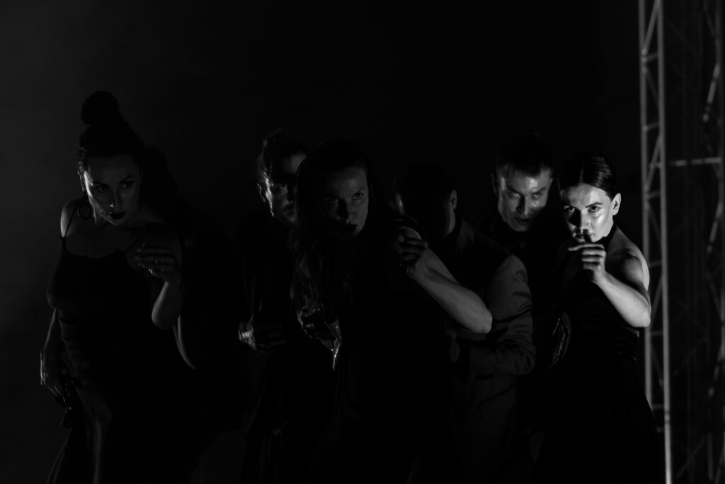 Zdjęcie z Tribute To, zdjęcie jest czarno białe. Na zdjęciu grupa tancerzy zarówno kobiet jak i mężczyzn, którzy wzrok maja skierowany w kierunku widowni i mają wyciągnięte do przodu lewe ręce.