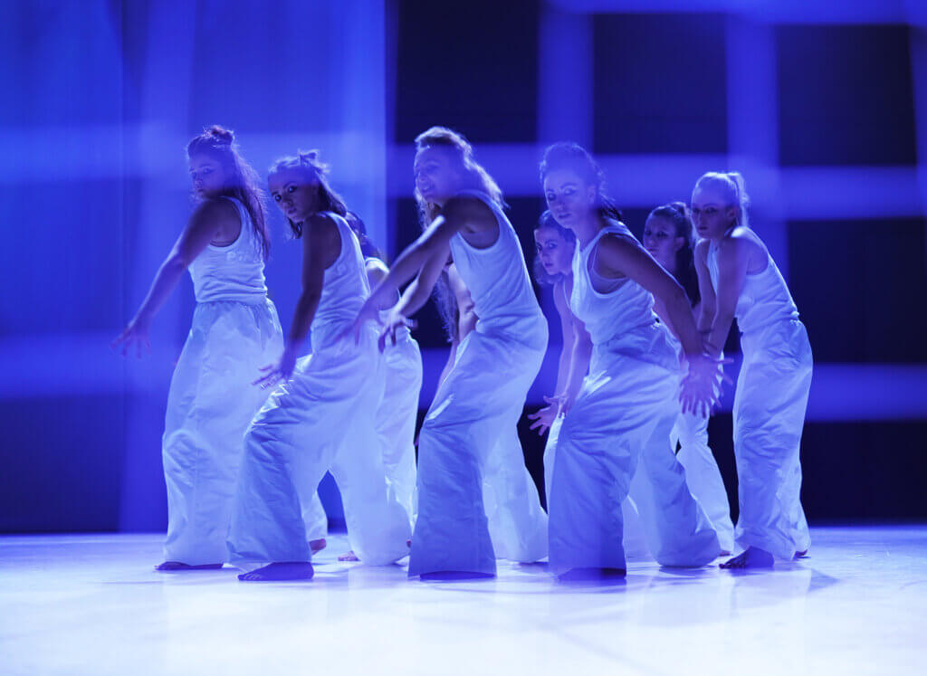 Zdjęcie ze spektaklu Dziadowisko. Na zdjęciu grupa kilku tancerek, ubranych w białe podkoszulki i białe spodnie. Tanerki przemieszczają się w prawą stronę sceny a głowy mają skierowane w lewą stronę.