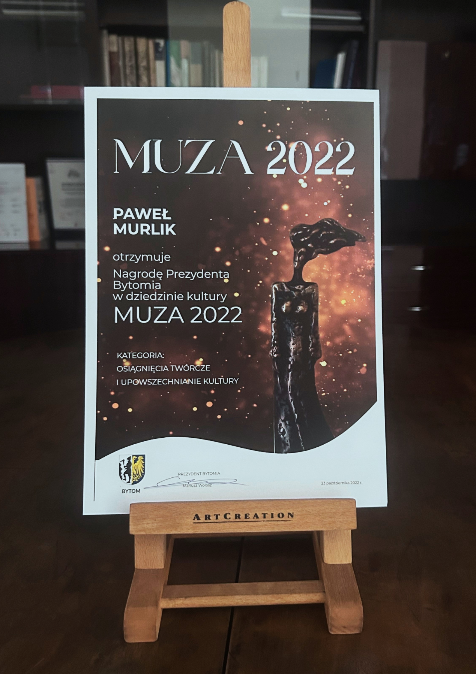 Plakat muza 2022. Z prawej strony statuetka kobiety z rozwianym włosem. Z lewej strony biały napis Paweł Murlik otrzymuje nagrodę Prezydenta Bytomia w dziedzinie kultury Muza 2022