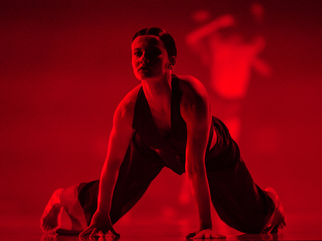 Zdjęcie ze spektaklu Bestie, na pierwszym planie jest tancerka która podpiera się rękami i stopami. Wzrok skierowany ma w stronę widowni, ubrana w czarny kostium.
