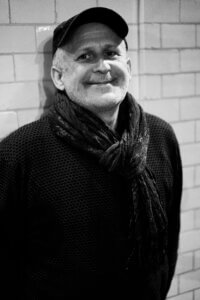 Zdjęcie czarno-białe na którym znajduje się uśmiechnięty mężczyzna, ubrany w ciemny swetr i ciemny szalik oraz czapkę z daszkiem. Mężczyzna stoi na tle białych cegieł.