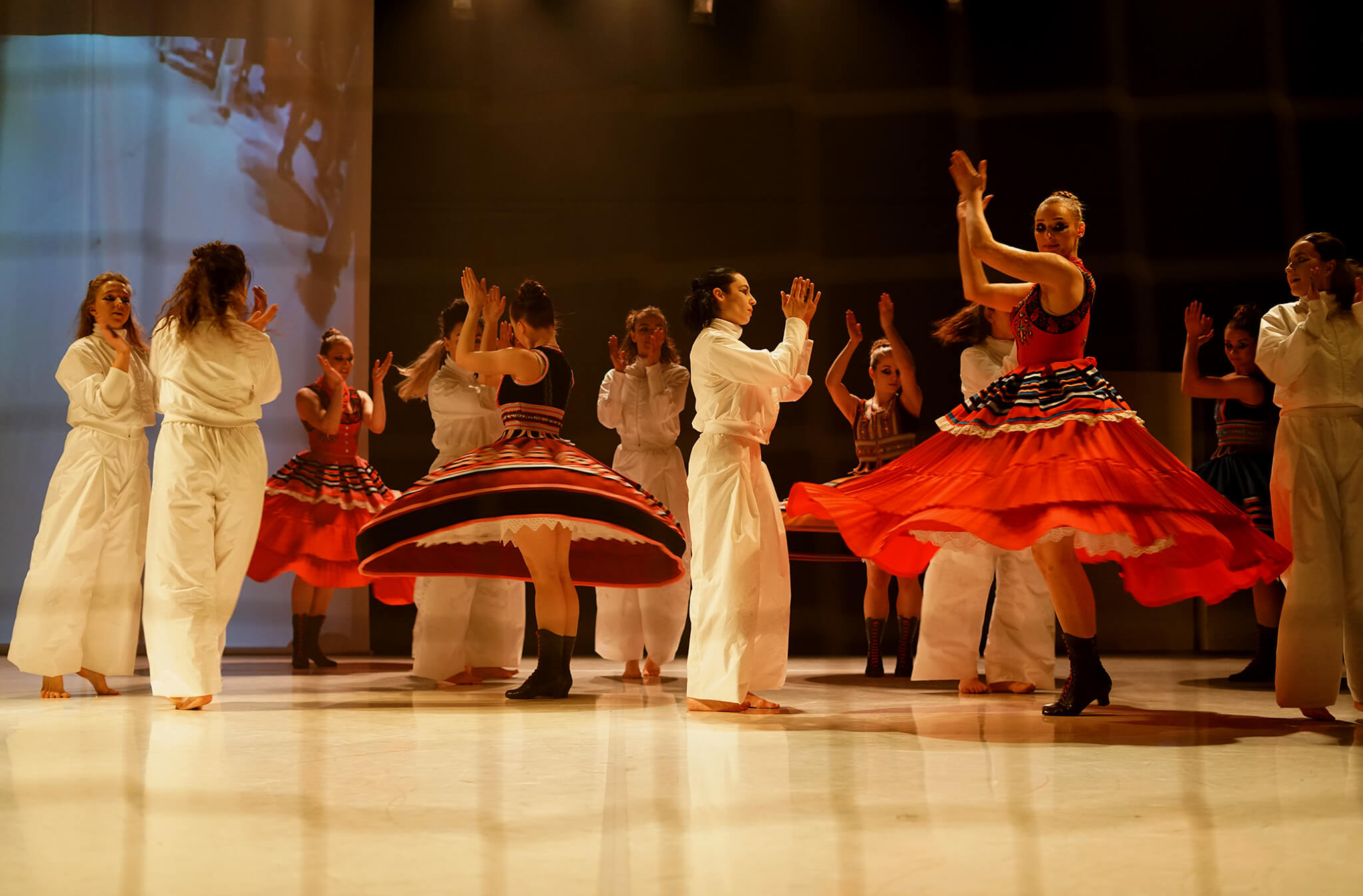zdjęcie ze spektaklu "DZIADOWISKO", kobiety w białych kombinezonach oraz inne w śląskich strojach ludowych, kręcą się na scenie i klaszczą w dłonie.
