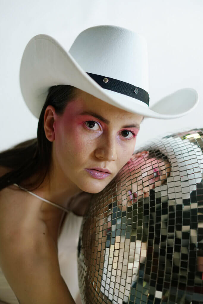 Zdjęcie z Fast Foot Fools na którym znajduje się młoda kobieta w białej koszulce i białym kowbojskim kapeluszu. Przytula się ona do srebrnej dyskotekowej kuli.