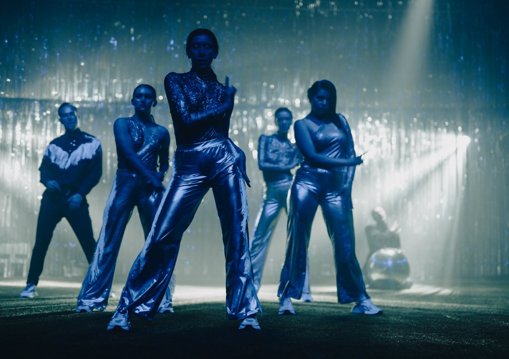 Zdjęcie ze spektaklu Fast Foot Fools, na zdjęciu znajduje się grupa tancerzy, stojących przodem do widowni, prawe ręce zgięte, ubrani są w srebrne kostiumy.