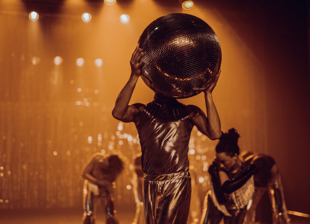 Zdjęcie ze spektaklu Fast Foot Fools, na zdjęciu znajduje się tancerz ubrany na srebrno, który na wysokości swojej twarzy trzyma dużą srebrną kule.