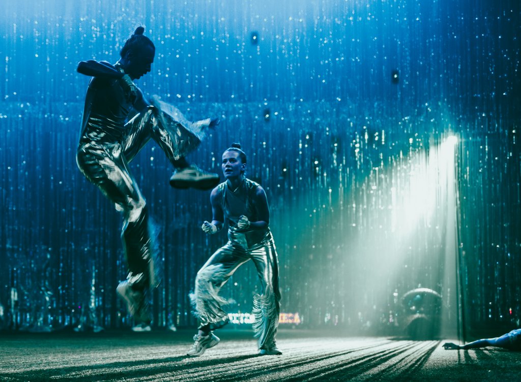 Zdjęcie ze spektaklu Fast Foot Fools. Na zdjęciu znajduje się dwoje tancerzy. jedena osoba skacze a druga stoi na ugiętych nogach.