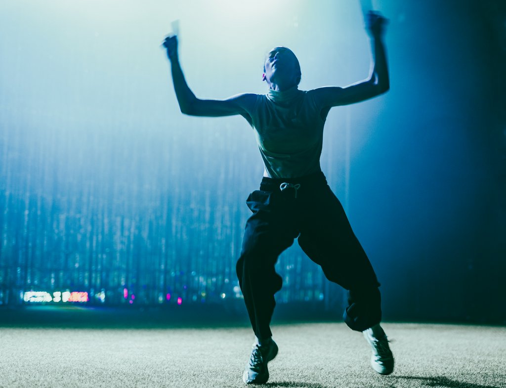 Zdjęcie ze spektaklu, na którym znajduje się tancerka stojąca w rozkroku, ręce ma uniesione w górę. Ubrana jest w golf bez rękawów, ciemne dresowe spodnie i buty sportowe.