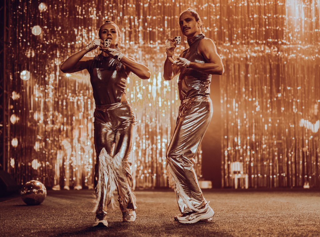 Zdjęcie ze spektaklu Fast Foot Fools. Na zdjęciu znajdują się dwie osoby mężczyzna i kobieta , oboje ubrani są w srebrne długi spodnie, srebrne topy i białe adidasy. Osoby stoją i trzymają malutkie filiżanki.