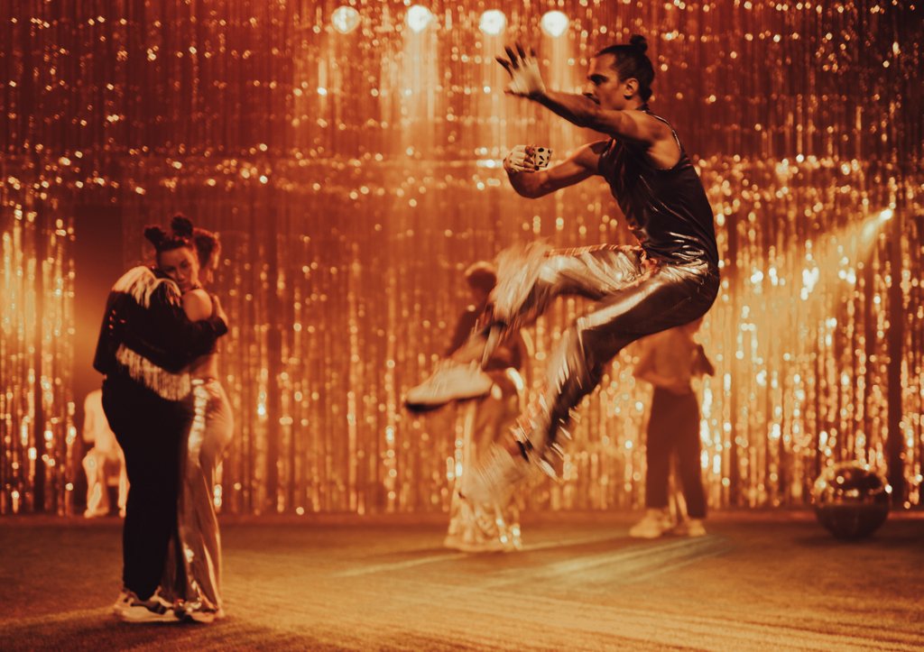 Zdjęcie ze spektaklu Fast Foot Fools. Na zdjęciu znajduje się kilku tancerzy na scenie, ubrani w srebrne stroje. Dwoje przytula się a jeden skacze.