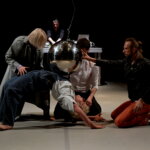 Zdjęcie ze spektaklu. Na srodku sceny na białej podłodze znajdują się cztery osoby. Dwie osoby klęczą, jedna stoi lekko pochylona, zas czwarta wykonuje mostek , starając się utrzymać na sobie dużą srebrną kulę.