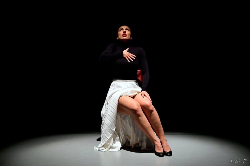 Zdjęcie ze spektaklu, na którym znajduje się tancerka siedząca na krześle. Ubrana jest w czarny golf i białą plisowaną spódnicę na stopach ma czarne szpilki.