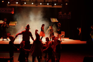 Zdjęcie ze spektaklu Bajlandia. Na zdjęciu znajduje się kilka osób tańczących w kole przed sceną.