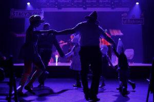 Zdjęcie ze spektaklu Bajlandia. Na zdjęciu znajduje się kilka osób tańczących w kole przed sceną.