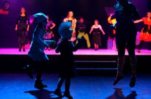 Zdjęcie ze spektaklu Bajlandia. Na zdjęciu znajduje się dwójka dzieci tańczaca przed sceną na głowie mają smerfowe czapki a w oddali na scenie znajduje się grupa aktorów tańcząca , ubrana w różnokolorowe bajkowe stroje.
