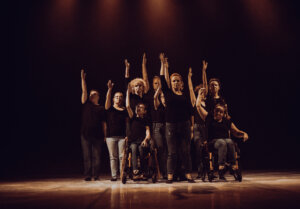 Zdjęcie ze spektaklu Integry. Na zdjęciu znajduje się grupa osób, stojąca twarzą do widowni i każda z tych osób ma uniesioną prawą rękę w górze. Wszyscy ubrani są w czarne koszulki i spodnie dżinsowe.