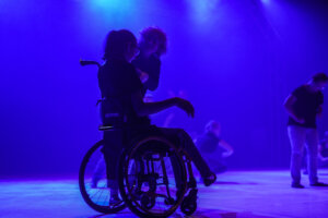 Zdjęcie zespektaklu Integry. Na zdjęciu znajduje się dziewczyna siedzaca na wózku inwalidzkim, ma lekko uniesioną rękę i nogę. Z jej lewej strony znajduje się kobieta, która trzyma ją za lewą rękę.