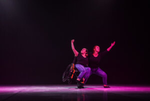 Zdjęcie ze spektaklu Integry. Na zdjęciu znajdują się dwie kobiety ubrane w czarne koszulki i spodnie dzinsowe. Jedna z nij jest na pomarańczowym wózku inwalidzkim. Osóba na wózku jest wsoarta na kolnie osby stojacej.