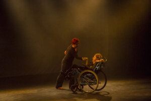 Zdjęcie ze spektaklu Integry. Na zdjęciu znajdują się dwie osoby. Jedna osoba jest na wózku inwalidzkim a druga jej asystuje w tańcu. Obie są ubrane na czarno.