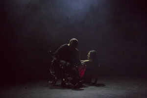 Zdjęcie Integry. Na zdjęciu znajduje się mężczyzna siedzący na wózku inwalidzkim, ubrany na czarno a obok niego na ziemi siedzi kobieta ubrana w czerwoną sukienkę.