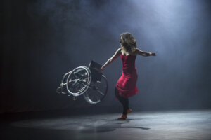 Zdjęcie integry. Na zdjęciu znajduje się kobieta ubrana w czerwoną sukienkę. Kobieta ma rozpuszczone włósy.W prawej ręce trzyma uniesiony pusty wózek inwalidzki, z którym wykonuje piruet.